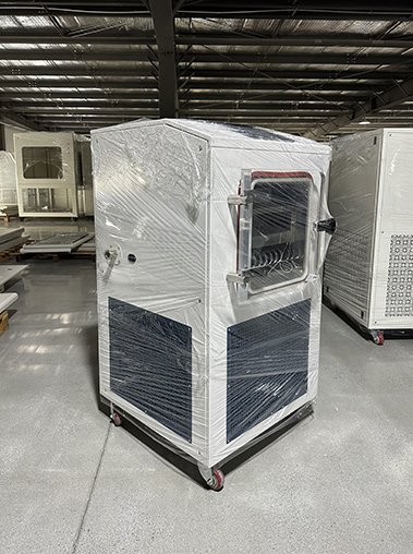 Front view of a white laboratory freeze dryer wrapped in protective plastic film on a warehouse factory floor.